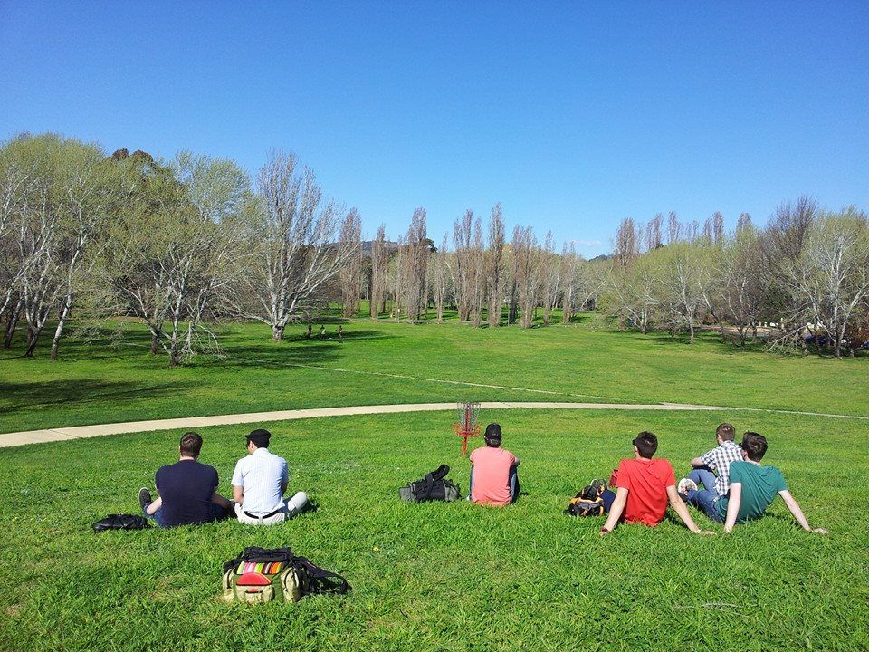 Disc golfers sitting on the grass beside a basket at Eddison Park Disc Golf Course in the Australian Capital Territory, overlooking open parkland and trees.