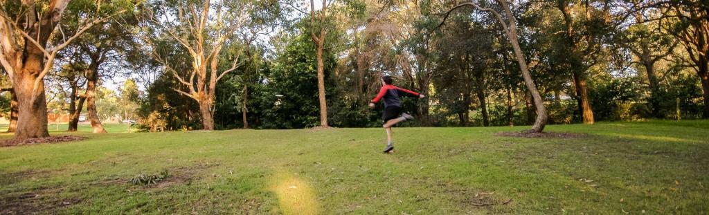 Disc golfer throwing across an open fairway at Jesmond Park Disc Golf Course, surrounded by mature trees and grassed parkland.