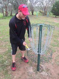 Player retrieving a disc from a basket at Meldanda Disc Golf Course, with native bushland and open grass in the background.