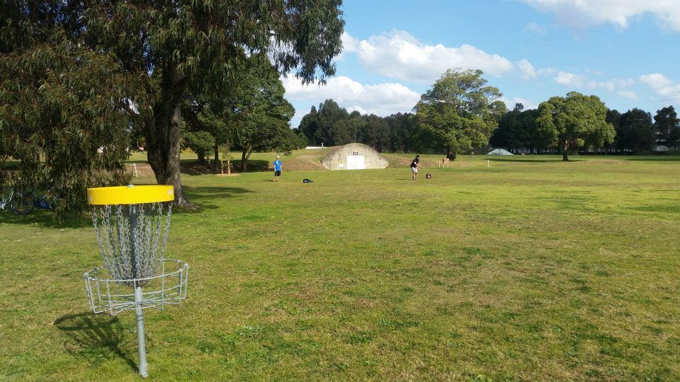 Newington Armoury Disc Golf Course with a basket in the foreground, open grass fairway and players throwing in the distance.