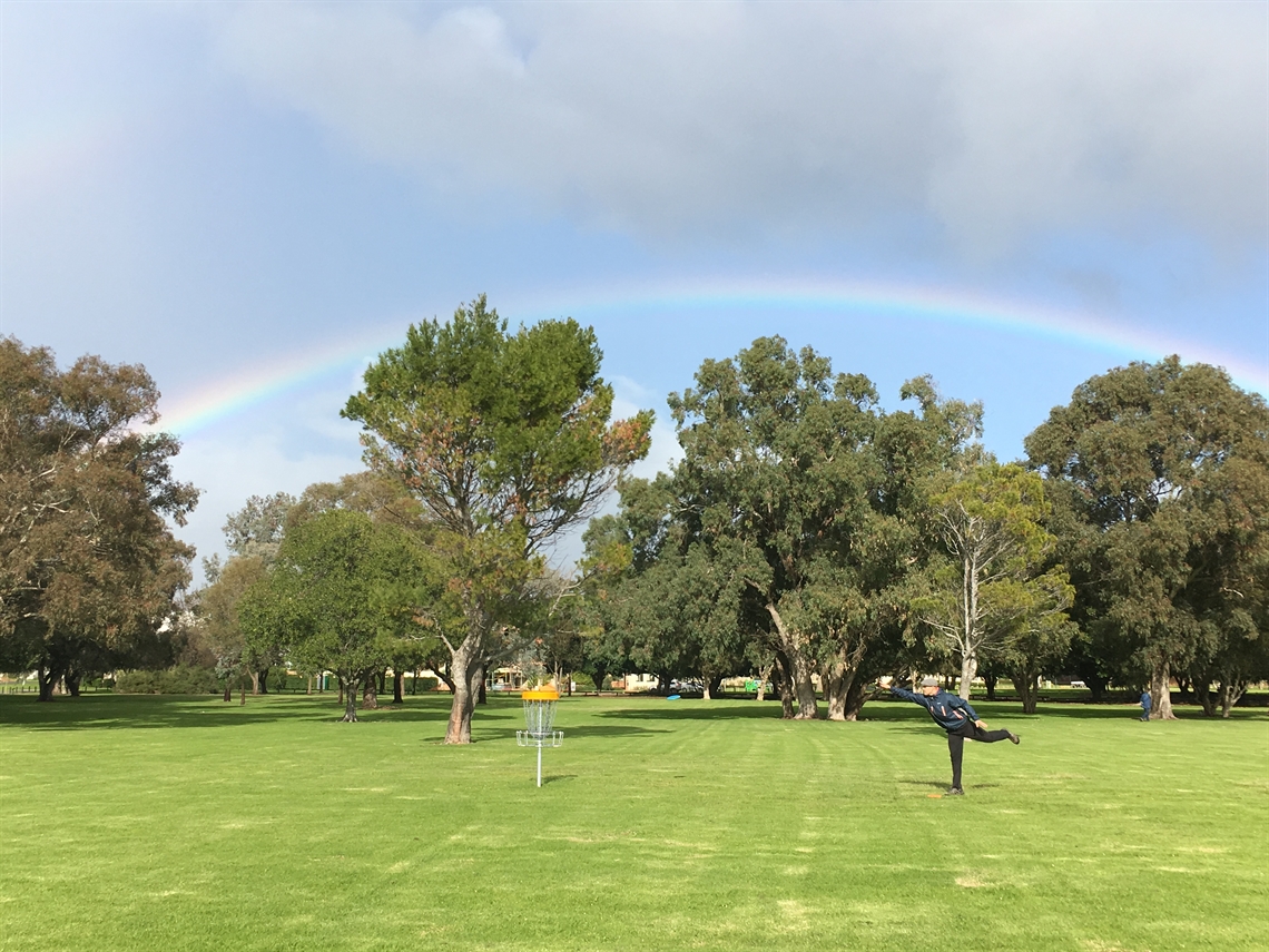 A player following through on a throw across open parkland, a rainbow arcing over the tree line.