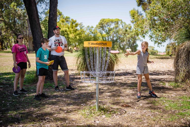 A family playing disc golf together in a park, a girl throwing towards the basket while the others watch.