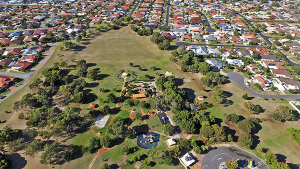 Aerial view of Fantasy Park, a suburban reserve ringed by houses, with open lawn, mature trees and a playground.