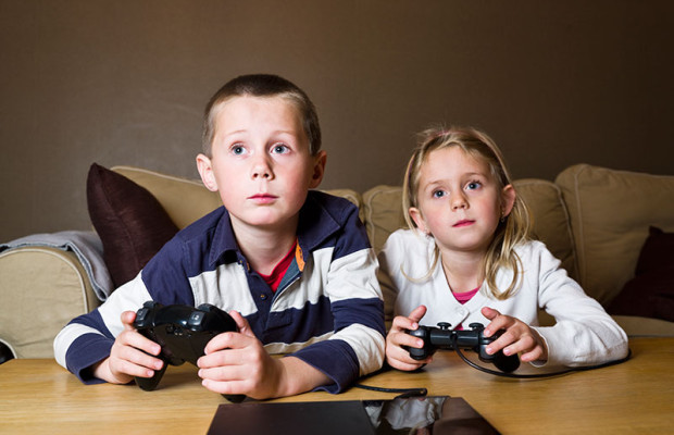 Two young children sitting on a sofa playing a video game with handheld controllers.
