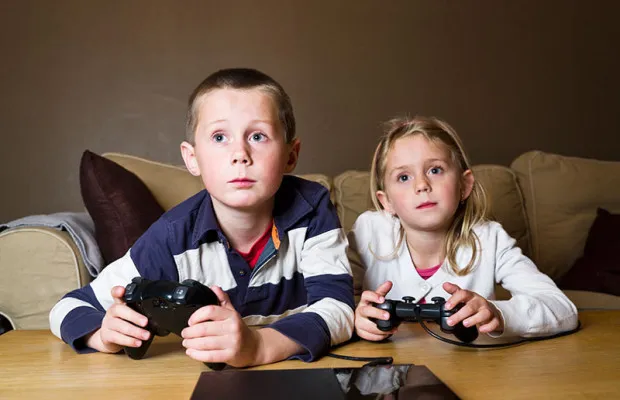 Two young children sitting on a sofa playing a video game with handheld controllers.