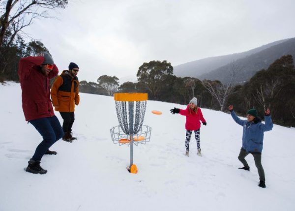A group playing disc golf in the snow around a basket on a winter alpine slope, throwing an orange disc.