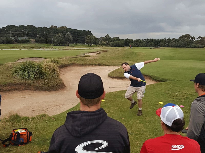 A player throwing a disc across a manicured golf course fairway while three spectators watch from beside a bunker.
