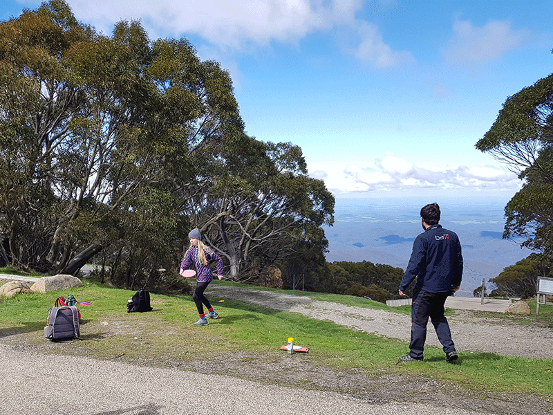Two players on a gravel path high on an alpine ridge, one carrying discs, with cloud filling the valley below.