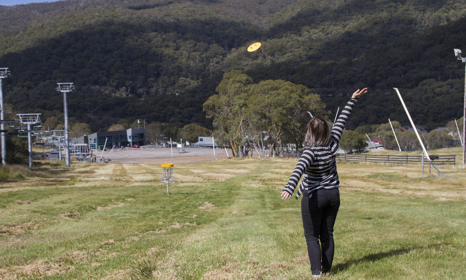 A woman throwing a disc uphill towards a ski village, the disc in mid-air above the slope.