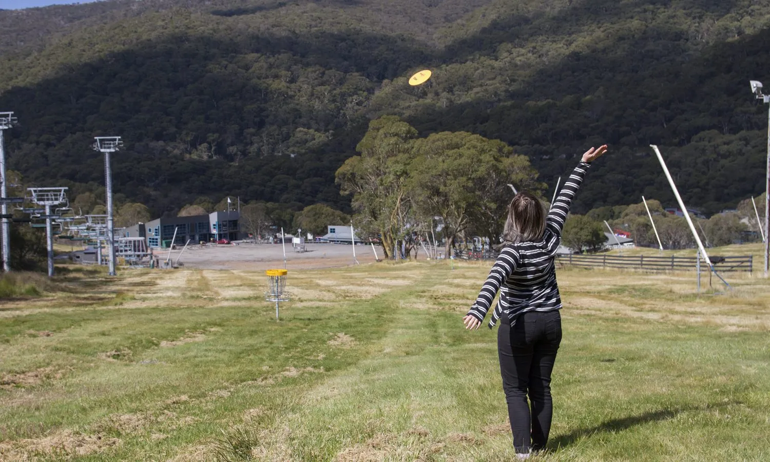A woman throwing a disc uphill towards a ski village, the disc in mid-air above the slope.