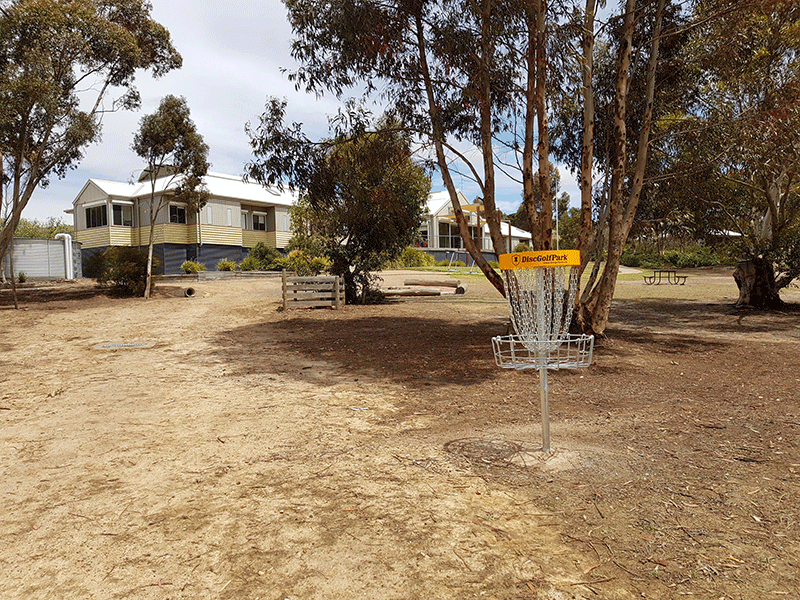 A disc golf basket on bare ground beside camp accommodation buildings and gum trees.