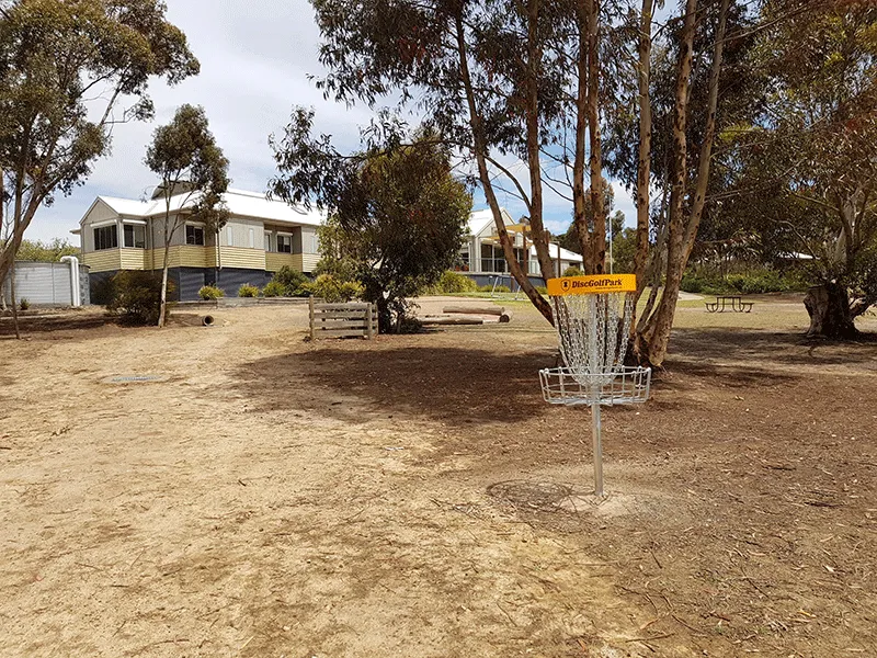 A disc golf basket on bare ground beside camp accommodation buildings and gum trees.