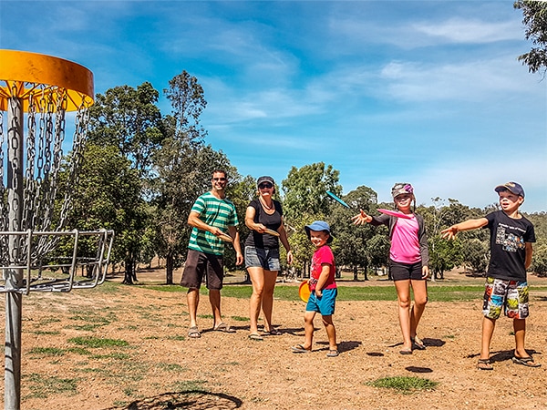 A family of five on a sandy course pointing and throwing towards a disc golf basket.