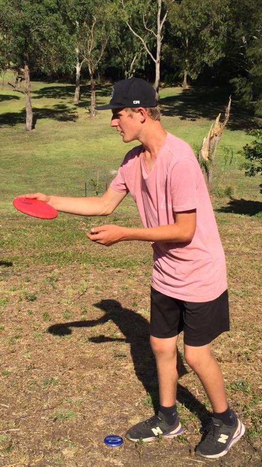A young player in a pink shirt holding a red disc, lining up a throw on a bushland course.