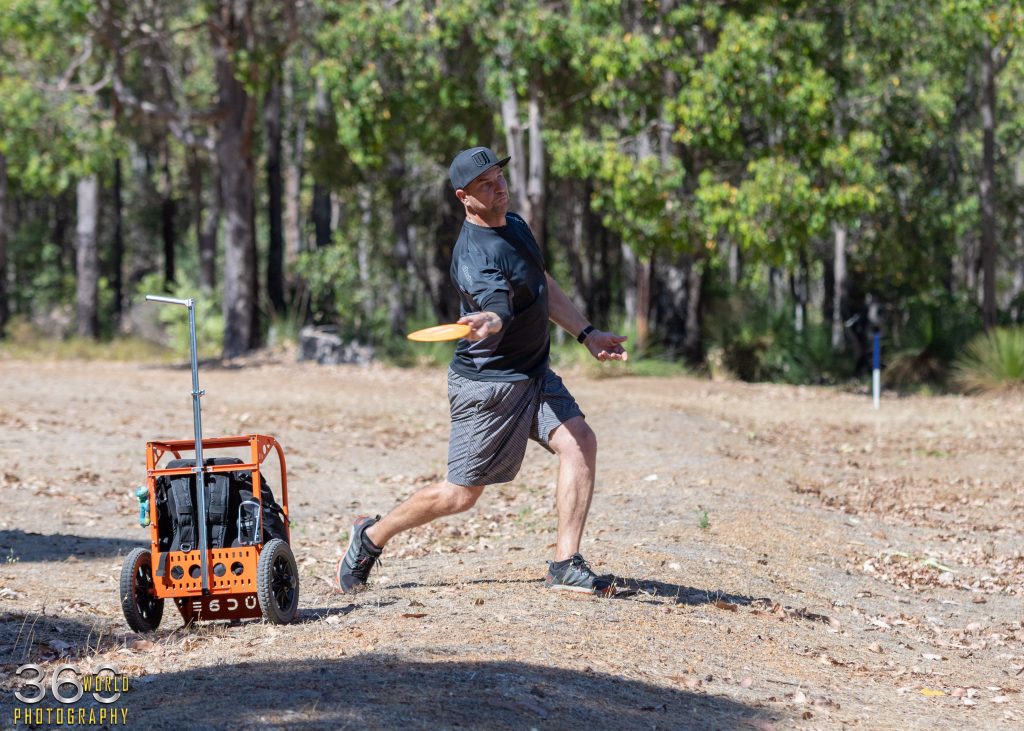A player mid-throw beside a wheeled disc golf cart on a dry, tree-lined course.