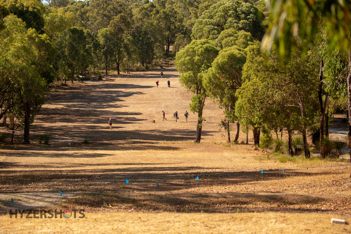 A long dry fairway lined with gum trees, players visible in the distance.