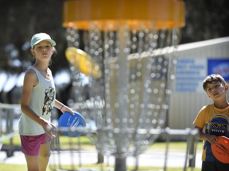 A girl and a boy holding discs beside a disc golf basket.