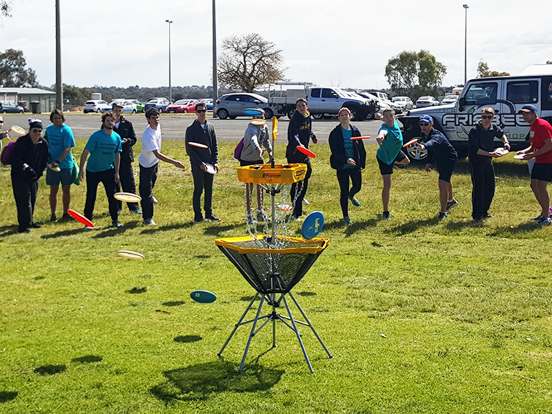 A group of players gathered around a disc golf basket on grass at a club come-and-try event.