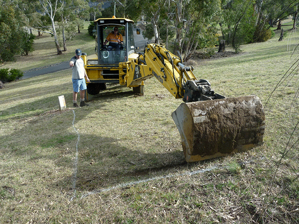 A yellow excavator lifting a log into place on grass while a worker looks on during course construction.
