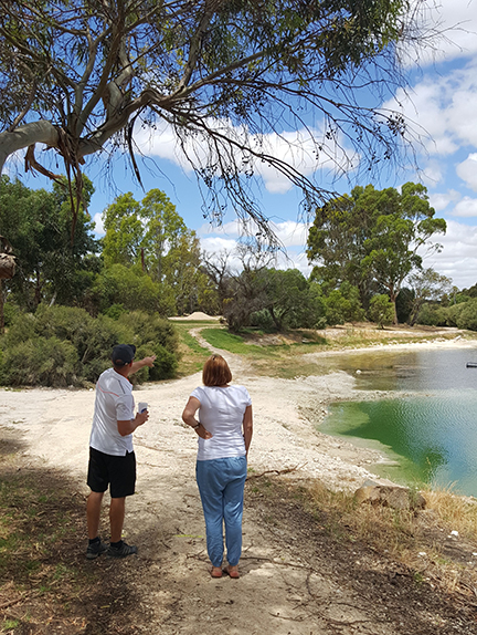 Two people standing on a dirt track beside a lake with bright green water, framed by a gum tree.
