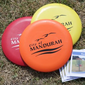 Three discs custom stamped with the City of Mandurah logo, resting on grass beside course flyers.