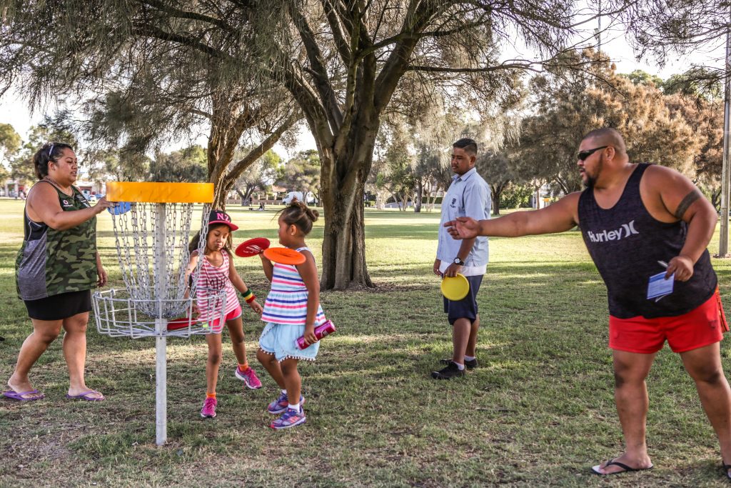 A family group of adults and children gathered with discs around a basket beneath a large shady tree.