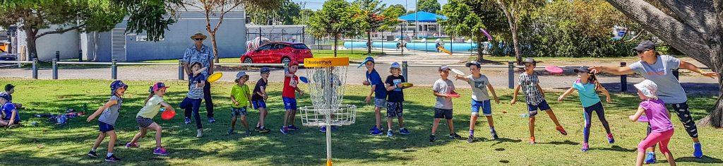 Children spread across a grassy reserve playing disc golf around a basket, beside a community pool.