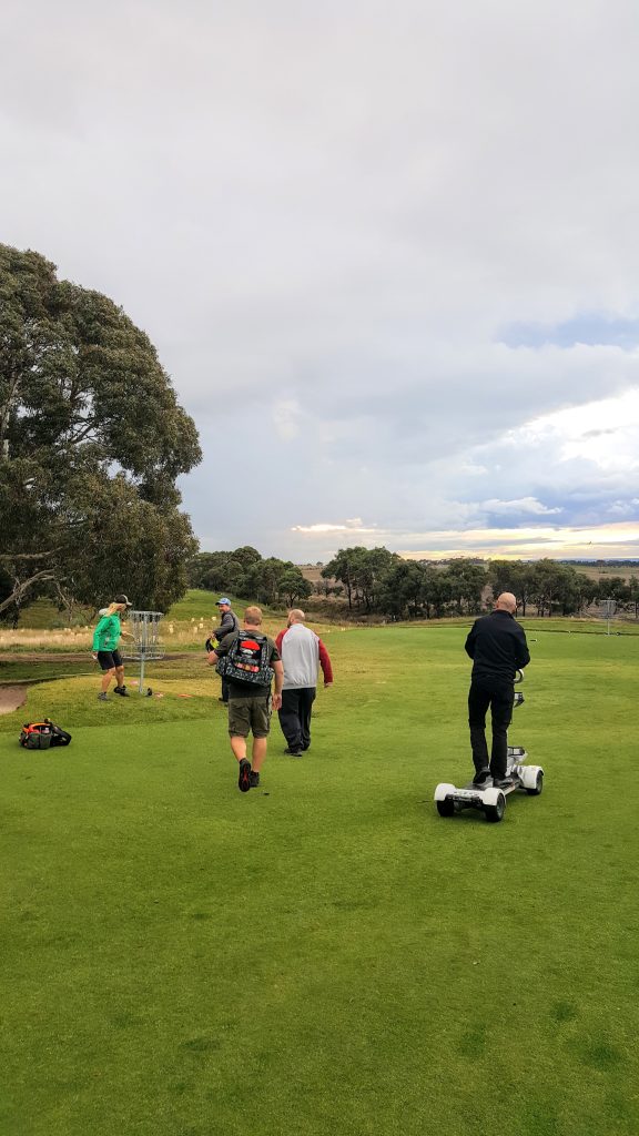 Players walking a golf course fairway with a storm building over the hills beyond.