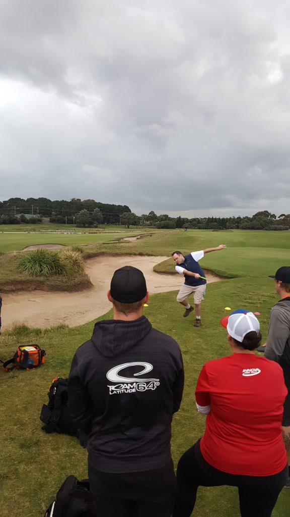 Spectators watching a player throw across a bunker on a golf course fairway.
