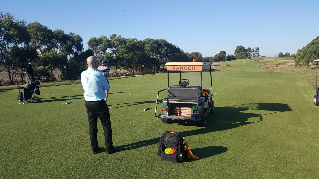 A man in business dress standing beside a golf course ranger cart on a manicured green.