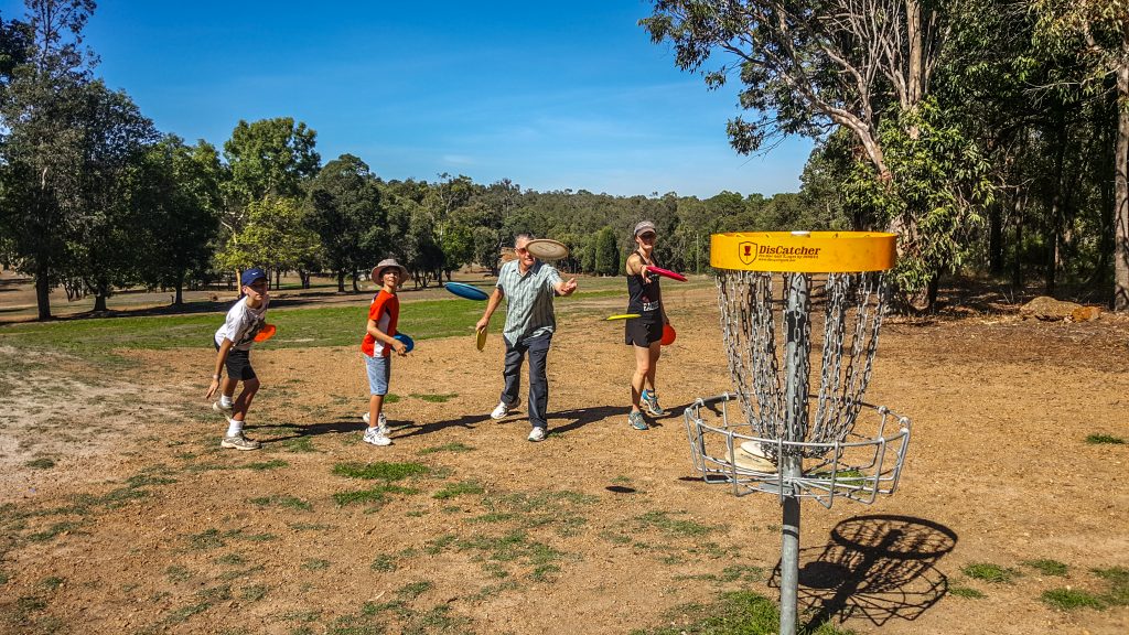 Children and adults gathered around a disc golf basket on a dry, tree-lined course.