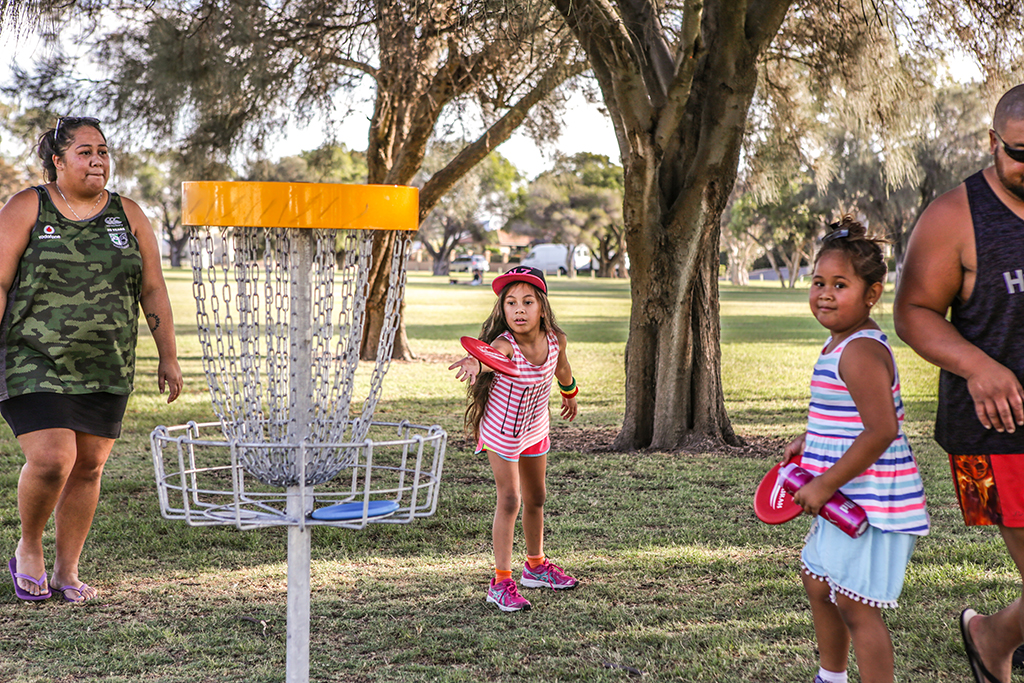 A family with young children gathered around a disc golf basket under a large tree.