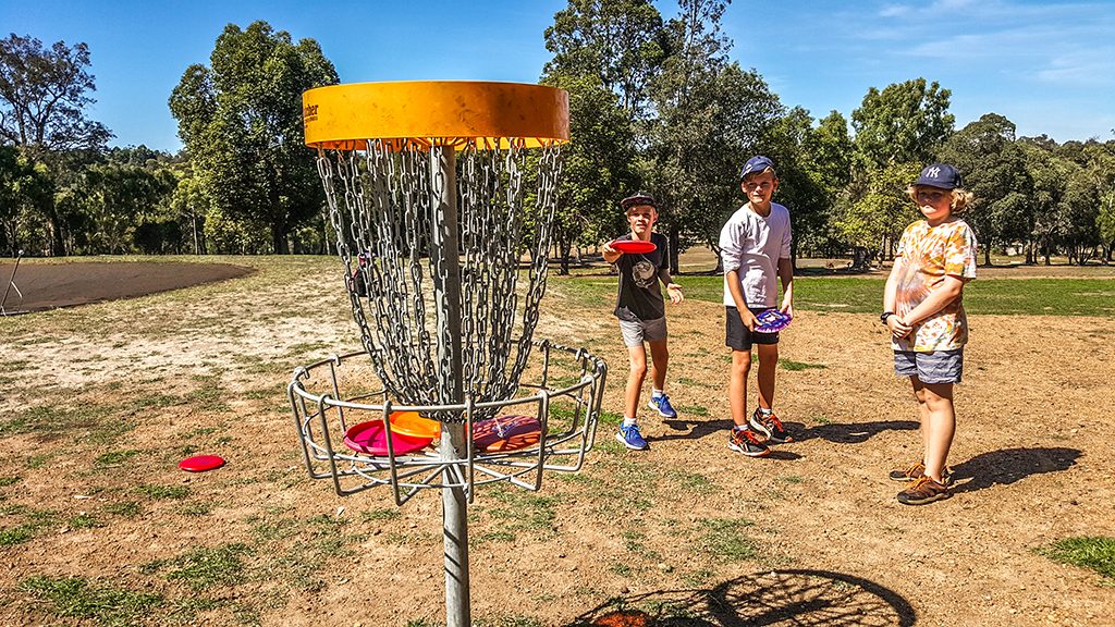 Three children standing around a disc golf basket on a dry course, holding coloured discs.