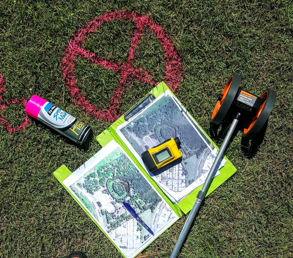A course design kit laid out on grass: marking spray, aerial site plans, a laser rangefinder and a measuring wheel, beside a target sprayed on the turf.