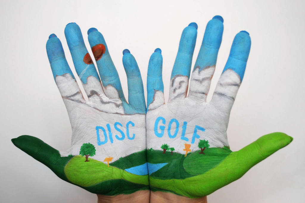 Two painted hands forming a landscape of sky, mountains and grass, with the words disc golf across the palms.