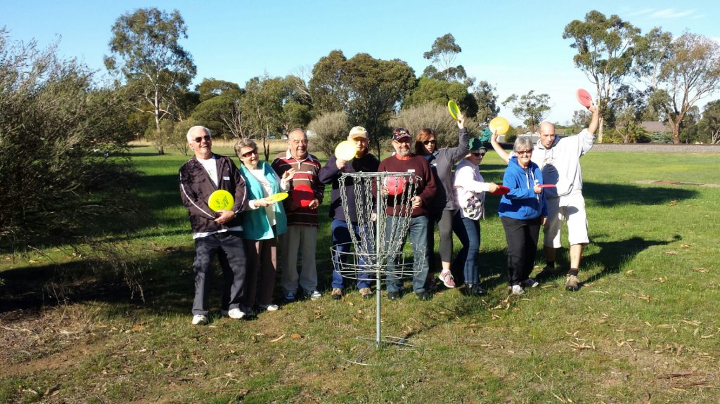 A group of adults gathered around a basket on a green fairway, several raising discs.