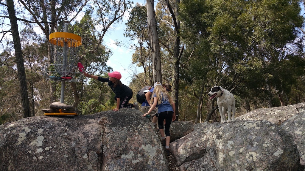 A disc golf basket set on a granite outcrop, players climbing the rocks towards it.