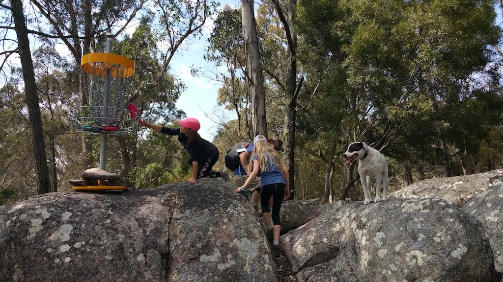 A disc golf basket set on a granite outcrop, players climbing the rocks towards it.