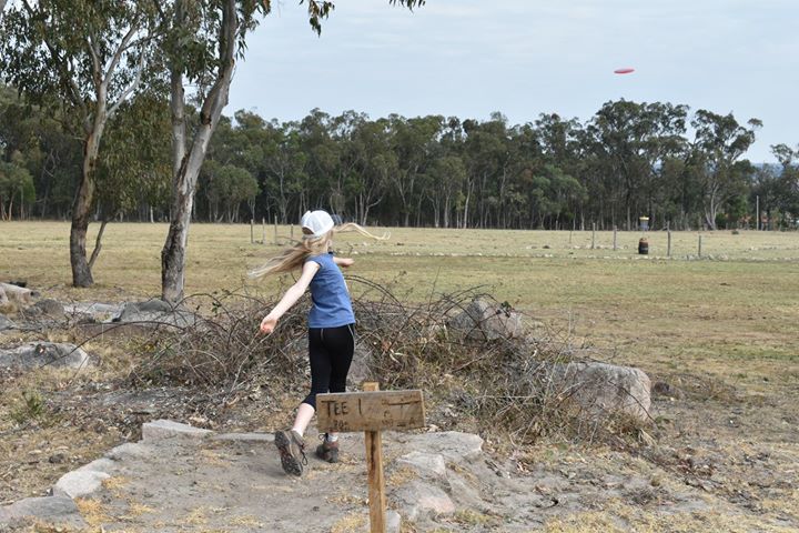 A player throwing from a rocky tee area in dry bushland, beside a Tee 1 sign.