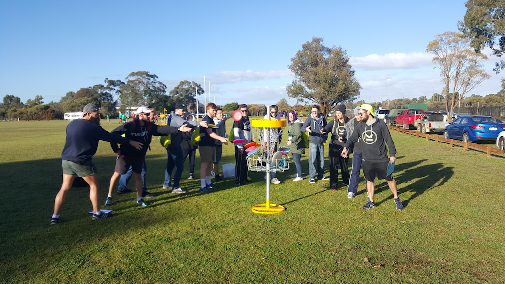 Adults gathered around a portable basket on grass at a community event, vehicles parked behind.