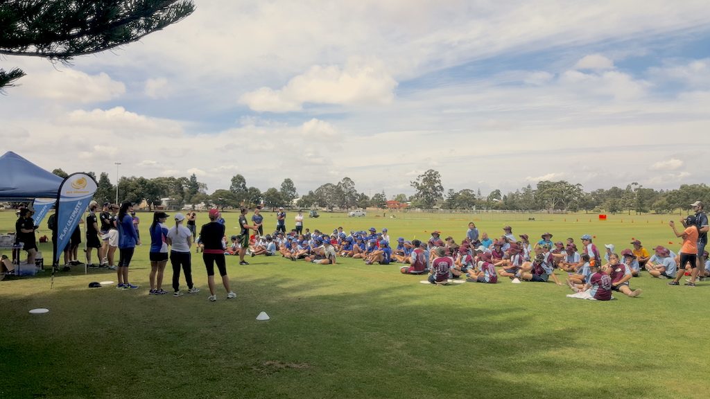 A large group of school children seated on an oval during a disc golf clinic, with instructors and a marquee.
