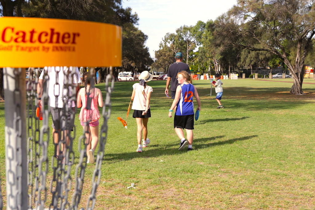 Close-up of a yellow DiscCatcher basket rim, with people walking on the grass beyond.