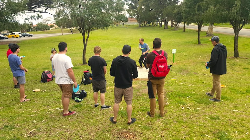 A group of adults standing on grass watching a player throw, bags resting beside them.