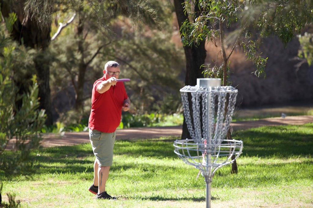 A player in a red shirt throwing towards a basket on a shaded parkland course.