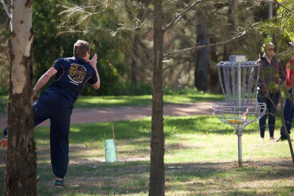 A player mid-throw among trees, a basket waiting on the fairway ahead.