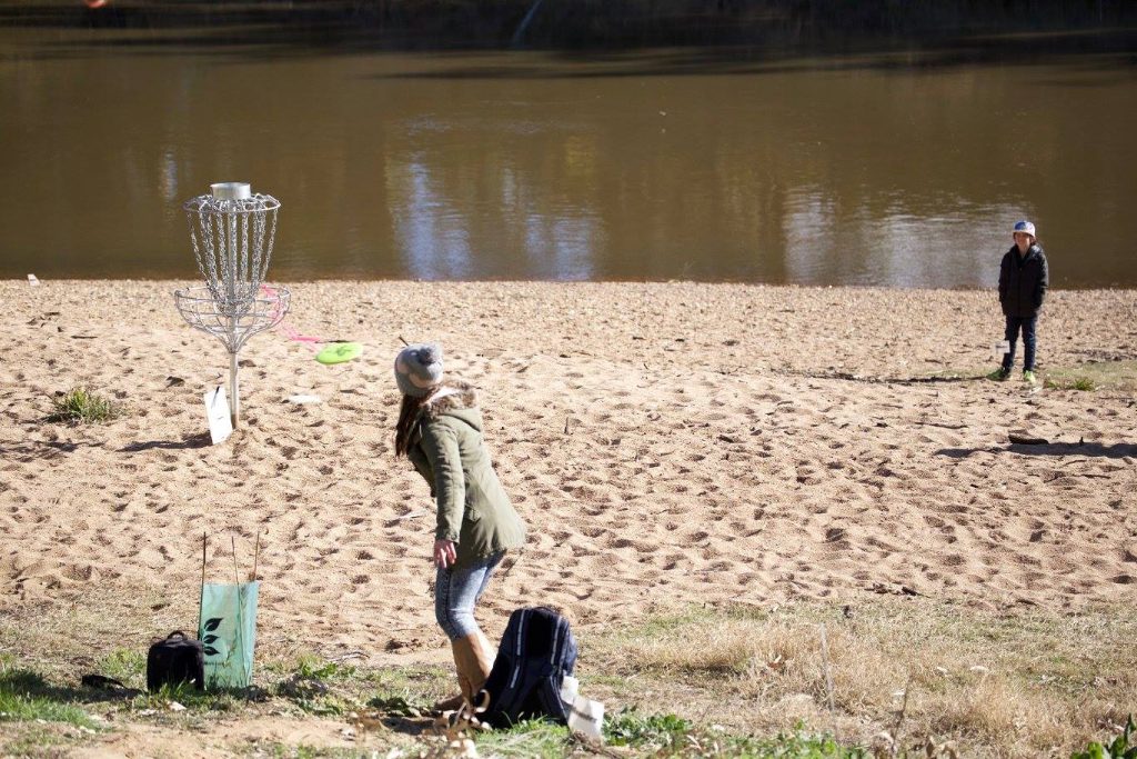 A player throwing on a sandy river beach, a disc golf basket standing at the water's edge.