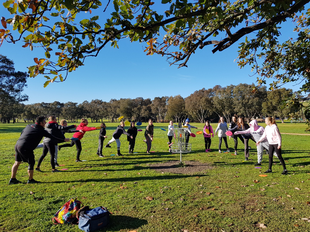 A large group of children and adults warming up together around a basket in a park.