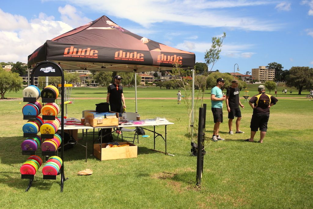 A branded event marquee with racks of disc golf bags and a merchandise table set up on grass.