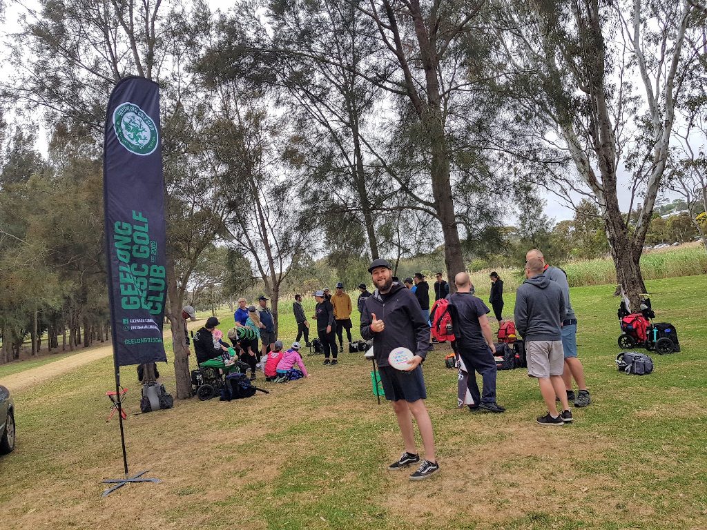 A Geelong Disc Golf feather flag beside players gathered on a grassy course.