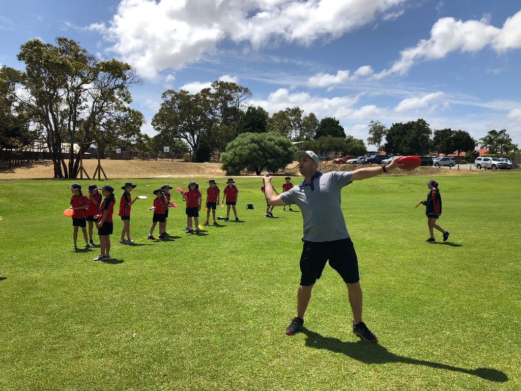 A coach throwing a disc on a green oval while school children in red uniforms watch.